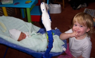Baby E in a bouncer with her big sister sitting next to her, looking very excited